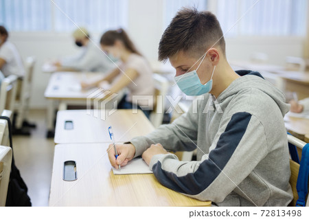 Teenager in protective mask writing in workbook on lesson Teenager in protective mask writing in workbook on lesson 72813498