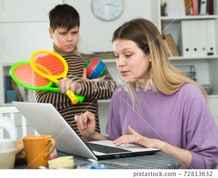 Mother working at laptop and unhappy son standing racquets for tennis Mother working at laptop and unhappy son standing racquets for tennis 72813632