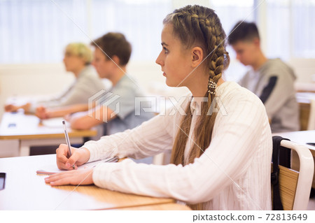 Teen girl listening to lecturer and writing in notebook in classroom Teen girl listening to lecturer and writing in notebook in classroom 72813649