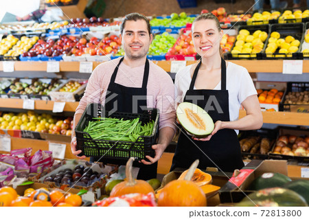 Young male seller standing with box of green beans and woman with half of melon on the supermarket Young male seller standing with box of green beans and woman with half of melon on the supermarket 72813800