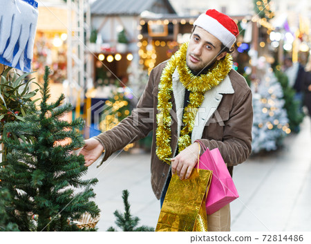 Young positive man buying Christmas tree and toys at Christmas Fair Young positive man buying Christmas tree and toys at Christmas Fair 72814486