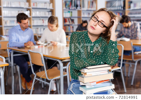 Annoyed girl sitting with pile of books in high school library on background of working fellow students Annoyed girl sitting with pile of books in high school library on background of working fellow students 72814991