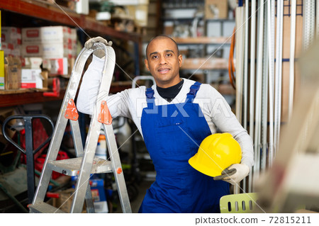 Positive warehouse worker stands next to stepladder and tool shelves in hardware store Positive warehouse worker stands next to stepladder and tool shelves in hardware store 72815211