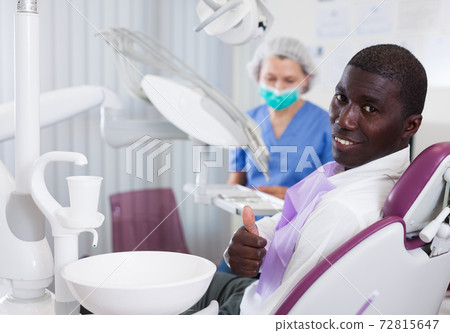 Happy african american man sitting in dental chair after teeth cure giving thumb up Happy african american man sitting in dental chair after teeth cure giving thumb up 72815647