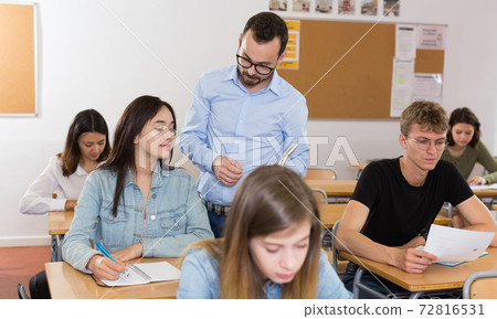 Smiling schoolgirl is writing answer the question on paper and teacher explaining it Smiling schoolgirl is writing answer the question on paper and teacher explaining it 72816531