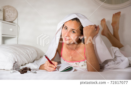 Young smiling woman writing to diary under blanket in bed Young smiling woman writing to diary under blanket in bed 72817077