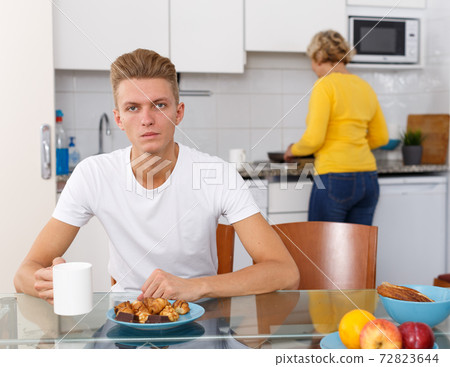 Guy sitting while his mother cooking breakfast Guy sitting while his mother cooking breakfast 72823644