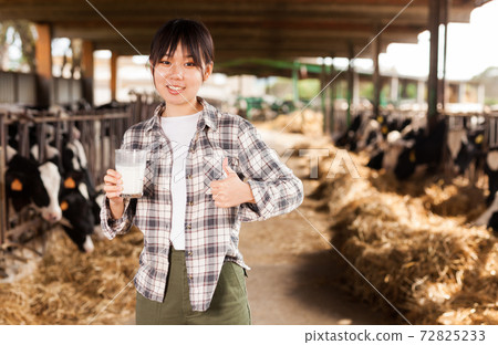 Young positive female dairy farm worker standing with glass of fresh milk Young positive female dairy farm worker standing with glass of fresh milk 72825233