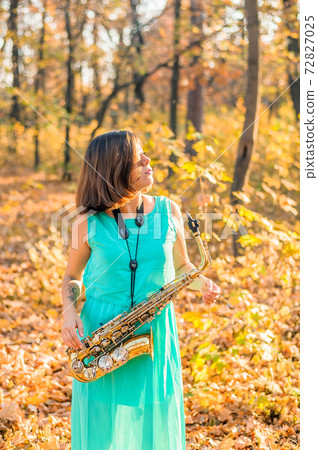 a girl with a beautiful figure and black hair looks away, standing in a yellow autumn park in a long blue dress with an alto saxophone in her hands a girl with a beautiful figure and black hair looks away, standing in a yellow autumn park in a long blue dress with an alto saxophone in her hands 72827025