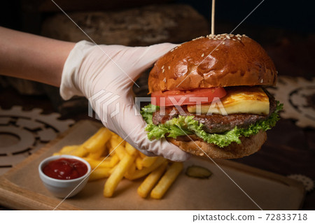 Hand in a disposable glow holding fresh craft beef burger with fried cheese, served with fresh lettuce, French fries and tomato sauce. Restaurant concept. Street food concept 72833718