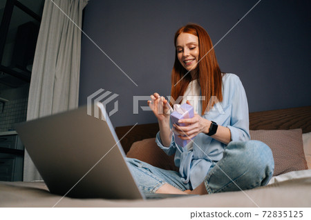 Low-angle shot of cheerful woman opening gift box with present while having video call on laptop. Low-angle shot of cheerful woman opening gift box with present while having video call on laptop. 72835125