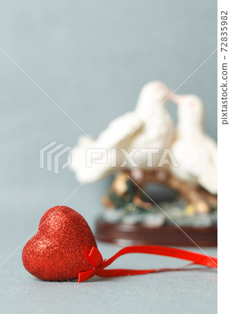 Red decorative shiny heart on a gray background. A statuette with kissing doves in the background. Copy space, selective focus Red decorative shiny heart on a gray background. A statuette with kissing doves in the background. Copy space, selective focus 72835982