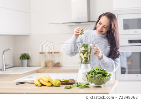 Woman standing in a kitchen squeezing lime into a blender full of fruit and vegetable Woman standing in a kitchen squeezing lime into a blender full of fruit and vegetable 72849368