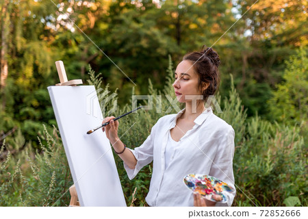 beautiful girl, woman artist, summer in park near the bushes, draws picture, palette with paints, brush white canvas painting, easel stand. White shirt. Green forest trees background. Hair in bun. 72852666