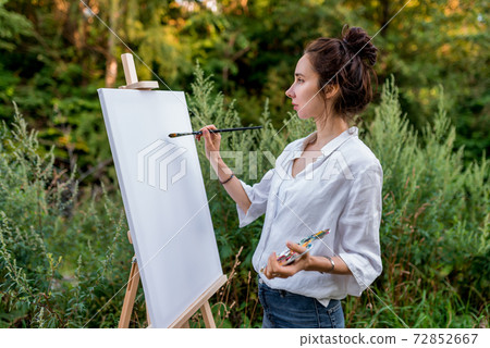 woman artist, in summer park by bushes, draws picture, palette paints, brush canvas painting, easel stand. White shirt. Green forest trees background. Creativity and idea creation. 72852667