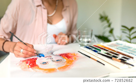 Close-up of female hands are painting a picture, a pink shirt at the artist, a portrait of a drawing girl, orange hair, a palette with flowers, a glass of water, brushes. Close-up of female hands are painting a picture, a pink shirt at the artist, a portrait of a drawing girl, orange hair, a palette with flowers, a glass of water, brushes. 72852682