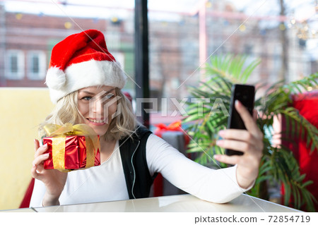 Celebrating Christmas in the distance. Smiling caucasian adult woman in Santa hat with red gift box in hand chatting with her family by smartphone in cafe, selective focus. 72854719