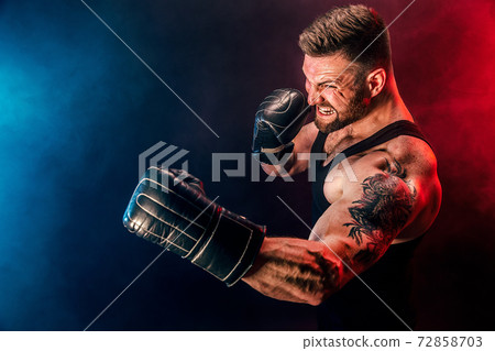Bearded tattooed sportsman muay thai boxer in black undershirt and boxing gloves fighting on dark background with smoke. 72858703