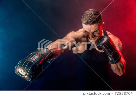 Bearded tattooed sportsman muay thai boxer in black undershirt and boxing gloves fighting on dark background with smoke. 72858704