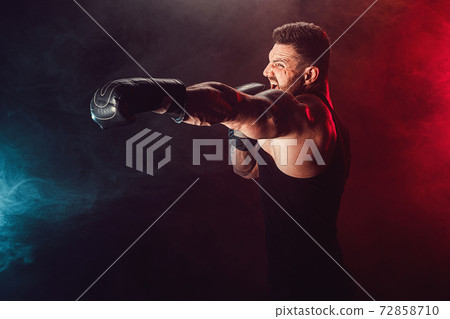 Bearded tattooed sportsman muay thai boxer in black undershirt and boxing gloves fighting on dark background with smoke. Bearded tattooed sportsman muay thai boxer in black undershirt and boxing gloves fighting on dark background with smoke. 72858710