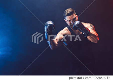 Bearded tattooed sportsman muay thai boxer in black undershirt and boxing gloves fighting on dark background with smoke. 72859212