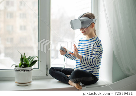 A child with a virtual reality headset is sitting at the table indoors at home. A girl exploring the world of virtual reality through video and games. 72859451