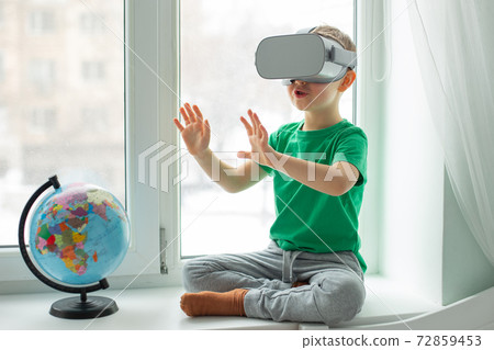 A boy with a virtual reality headset is sitting at home at the table indoors. The child explores the world of virtual reality through video and games. A boy with a virtual reality headset is sitting at home at the table indoors. The child explores the world of virtual reality through video and games. 72859453