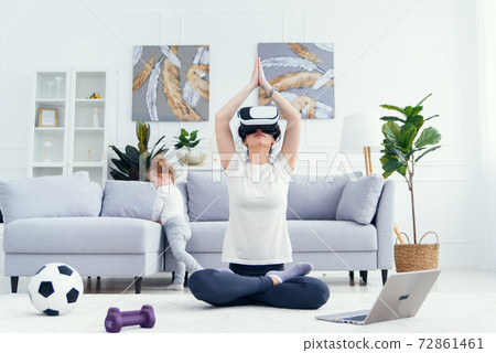 Young mom meditating in lotus yoga position using ar glasses while her daughter watches cartoons on background. Young mom meditating in lotus yoga position using ar glasses while her daughter watches cartoons on background. 72861461