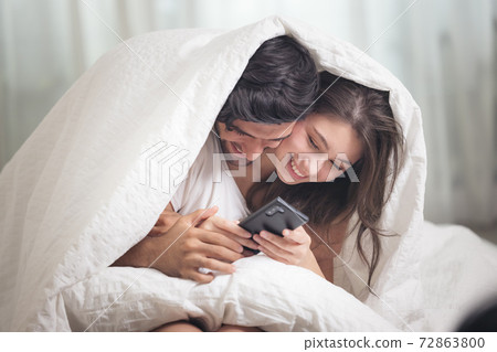 Young couple in bed, Happy smiling couple lying covered with blanket and Mobile Phone 72863800