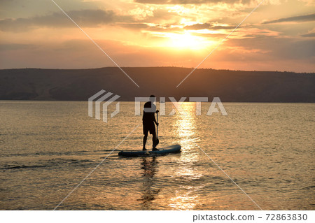 Man sail on a SUP board in a large lake during sunrise. Stand up paddle boarding active recreation in nature Man sail on a SUP board in a large lake during sunrise. Stand up paddle boarding active recreation in nature 72863830