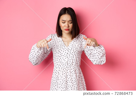 Concerned asian woman peeking down and pointing hand at disturbing banner, frowning upset, standing over pink background Concerned asian woman peeking down and pointing hand at disturbing banner, frowning upset, standing over pink background 72868473
