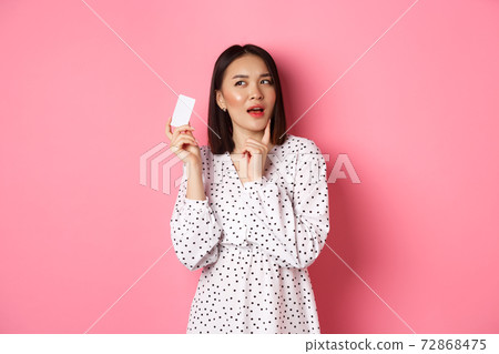 Shopping concept. Attractive asian woman thinking about buying presents, holding credit card and pondering, standing in dress against pink background Shopping concept. Attractive asian woman thinking about buying presents, holding credit card and pondering, standing in dress against pink background 72868475