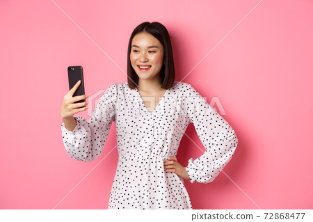 Beautiful asian girl using photo filters app and taking selfie on smartphone, posing in cute dress against pink background Beautiful asian girl using photo filters app and taking selfie on smartphone, posing in cute dress against pink background 72868477