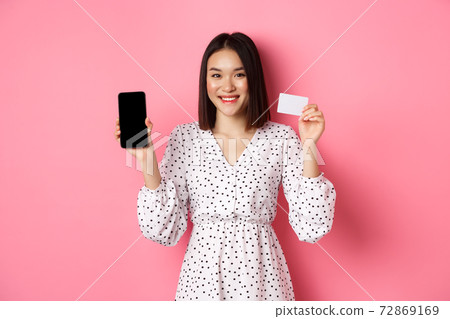 Cute asian woman shopping online, showing bank credit card and mobile screen, smiling and looking at camera, standing over pink background 72869169