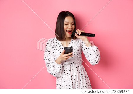 Cute asian woman reading lyrics in smartphone, singing with microphone, standing in trendy dress over pink background 72869275
