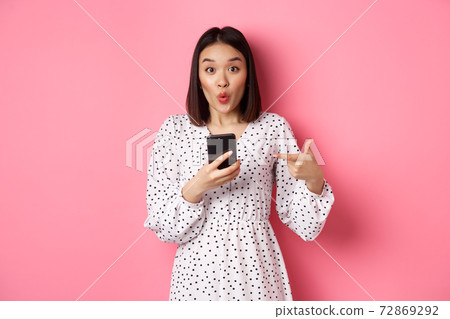 Online shopping and beauty concept. Amazed asian woman pointing finger at mobile phone, recommending smartphone app, standing over pink background 72869292