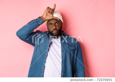 Sad african-american man showing loser sign on forehead and staring at camera, standing over pink background Sad african-american man showing loser sign on forehead and staring at camera, standing over pink background 72870091