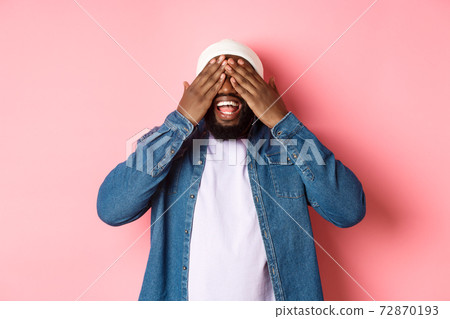 Happy Black man waiting for surprise, cover eyes and smiling, playing peekaboo, standing over pink background 72870193
