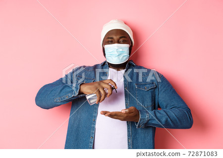 Covid-19, lifestyle and lockdown concept. Smiling african-american man in face mask cleaning hands with sanitizer, using antiseptic and looking at camera, pink background Covid-19, lifestyle and lockdown concept. Smiling african-american man in face mask cleaning hands with sanitizer, using antiseptic and looking at camera, pink background 72871083