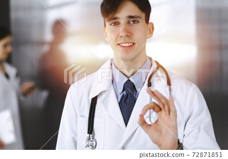 Friendly man-doctor standing straight and showing Ok sign in clinic. Female colleague is in a hurry at the background of physician. Medicine concept 72871851