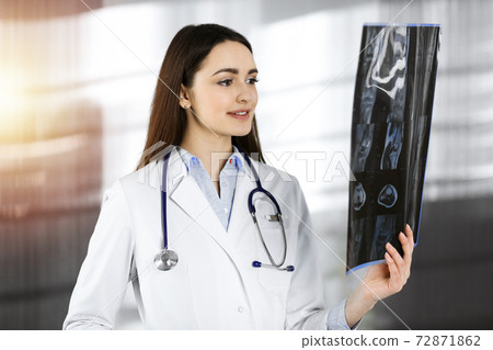 Professional young woman-doctor is looking at her patient's roentgenogram. Physician at work in sunny clinic Professional young woman-doctor is looking at her patient's roentgenogram. Physician at work in sunny clinic 72871862