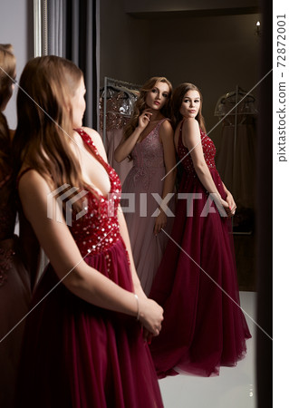 Two young beautiful girls wearing a full-length dark crimson red chiffon prom ball gown decorated with sparkles and sequins. Models in front of mirror in a fitting room at dress hire service. Two young beautiful girls wearing a full-length dark crimson red chiffon prom ball gown decorated with sparkles and sequins. Models in front of mirror in a fitting room at dress hire service. 72872001