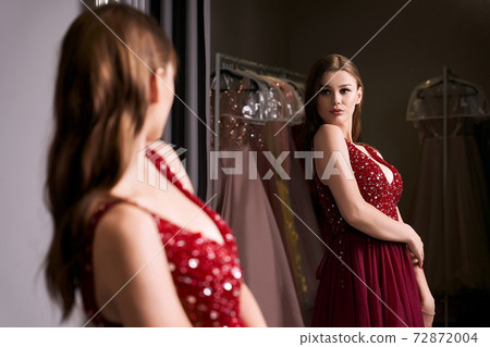 Young beautiful brunette girl wearing a full-length dark crimson red chiffon prom ball gown decorated with sparkles and sequins. Model in front of mirror in a fitting room at dress hire service. Young beautiful brunette girl wearing a full-length dark crimson red chiffon prom ball gown decorated with sparkles and sequins. Model in front of mirror in a fitting room at dress hire service. 72872004