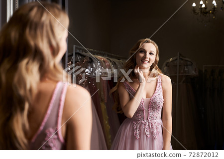 Young beautiful blonde girl wearing a full-length pink violet or purple chiffon prom ball gown decorated with sparkles and sequins. Model in front of mirror in a fitting room at dress hire service. Young beautiful blonde girl wearing a full-length pink violet or purple chiffon prom ball gown decorated with sparkles and sequins. Model in front of mirror in a fitting room at dress hire service. 72872012