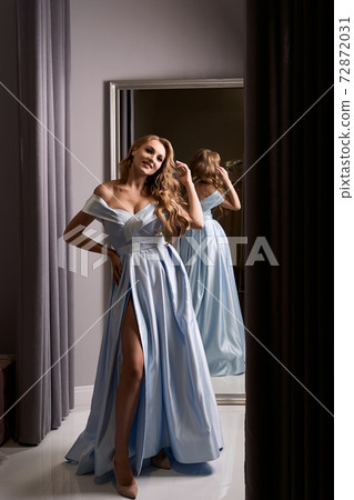 Young beautiful blonde girl wearing an off-the-shoulder full-length sky blue satin slit prom ball gown. Model looking in mirror. Fitting room in dress hire service. Young beautiful blonde girl wearing an off-the-shoulder full-length sky blue satin slit prom ball gown. Model looking in mirror. Fitting room in dress hire service. 72872031