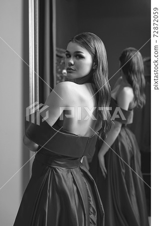 Young beautiful girl wearing an off-the-shoulder full-length satin slit prom ball gown. Model looking in mirror. Black and white image in retro style. Young beautiful girl wearing an off-the-shoulder full-length satin slit prom ball gown. Model looking in mirror. Black and white image in retro style. 72872059