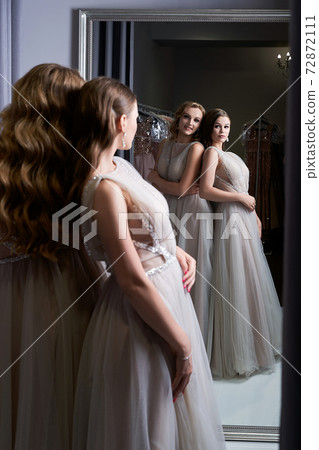 Two young beautiful girls wearing a full-length silver white chiffon prom ball gowns decorated with sparkles and sequins. Models in front of mirror in a fitting room. 72872111