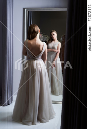 Young beautiful brunette girl wearing a full-length silver white chiffon prom ball gown decorated with sparkles and sequins. Model in front of mirror in a fitting room at dress hire service. 72872151