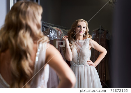 Young beautiful blonde girl wearing a full-length silver white chiffon prom ball gown decorated with sparkles and sequins. Model in front of mirror in a fitting room at dress hire service. Young beautiful blonde girl wearing a full-length silver white chiffon prom ball gown decorated with sparkles and sequins. Model in front of mirror in a fitting room at dress hire service. 72872165