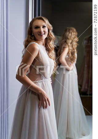 Young beautiful blonde girl wearing a full-length silver white chiffon prom ball gown decorated with sparkles and sequins. Model in front of mirror in a fitting room at dress hire service. 72872199
